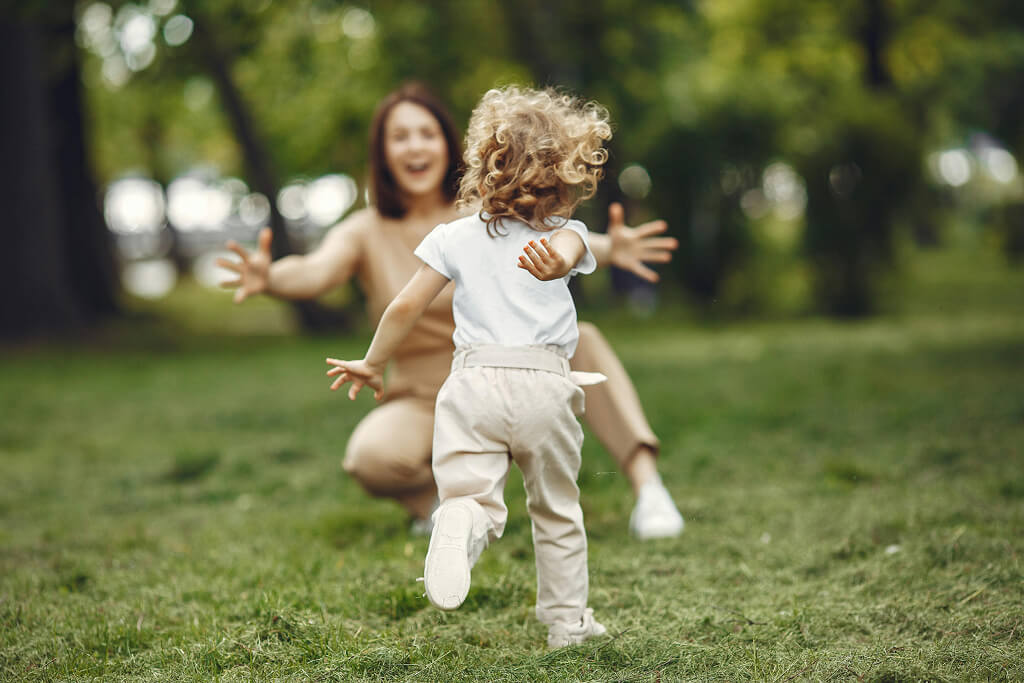 Child running with arms outstretched towards a crouching woman waiting with open arms in a park.