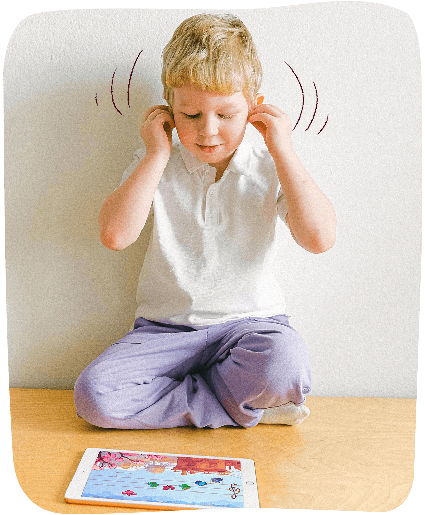 Young boy sitting cross-legged on a table with fingers in his ears, looking at a tablet displaying Elody game.