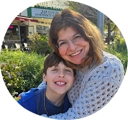 Smiling woman with glasses hugging a young boy outdoors with greenery in the background.