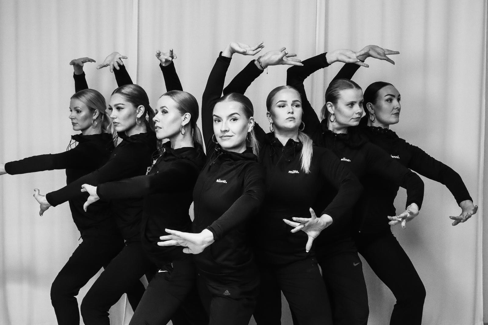 Seven women in black outfits striking a synchronized dance pose with expressive hand gestures.