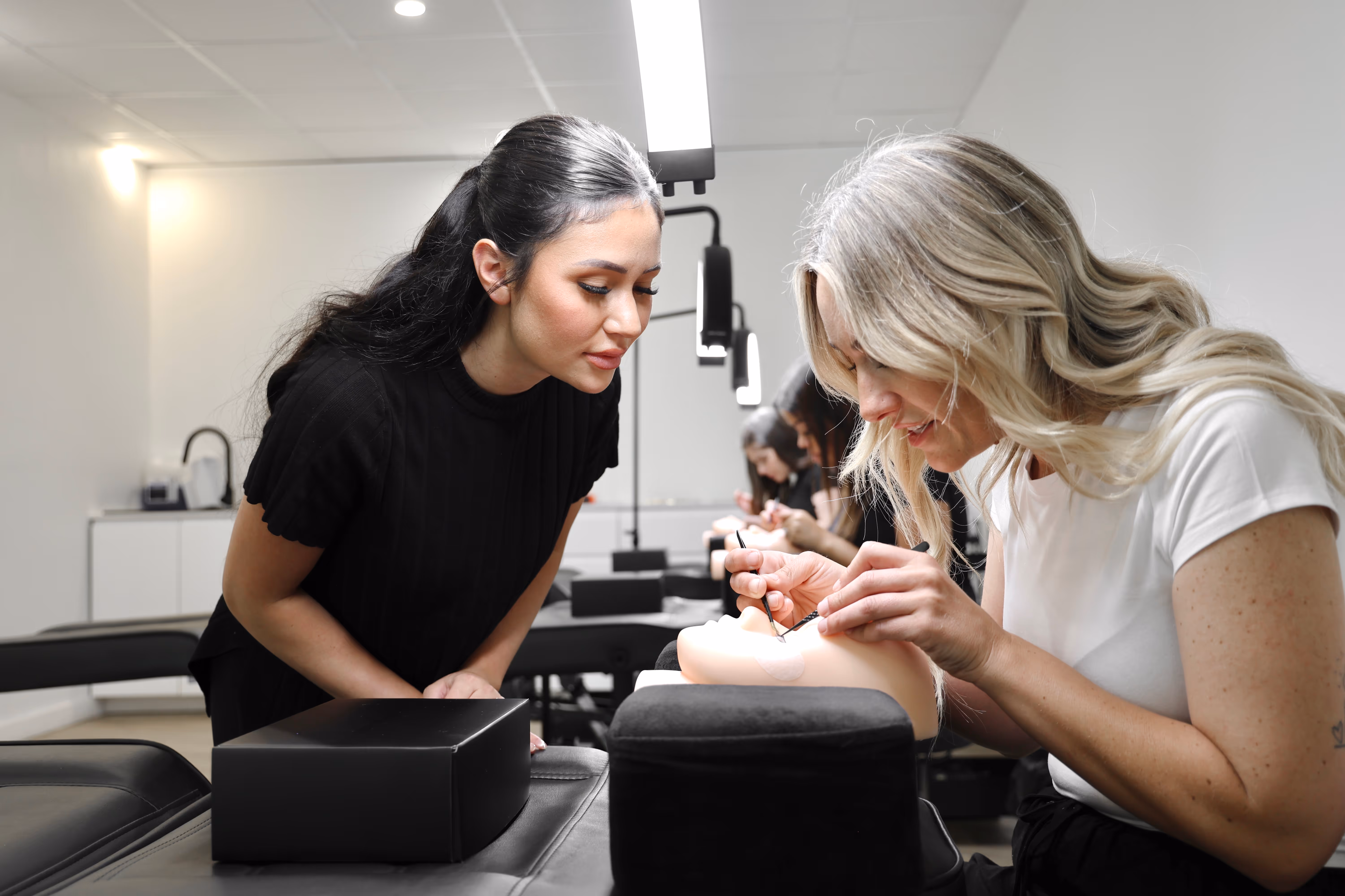 A blonde woman applying eyelash extensions to a practice mannequin while a dark-haired woman watches closely in a beauty training classroom.