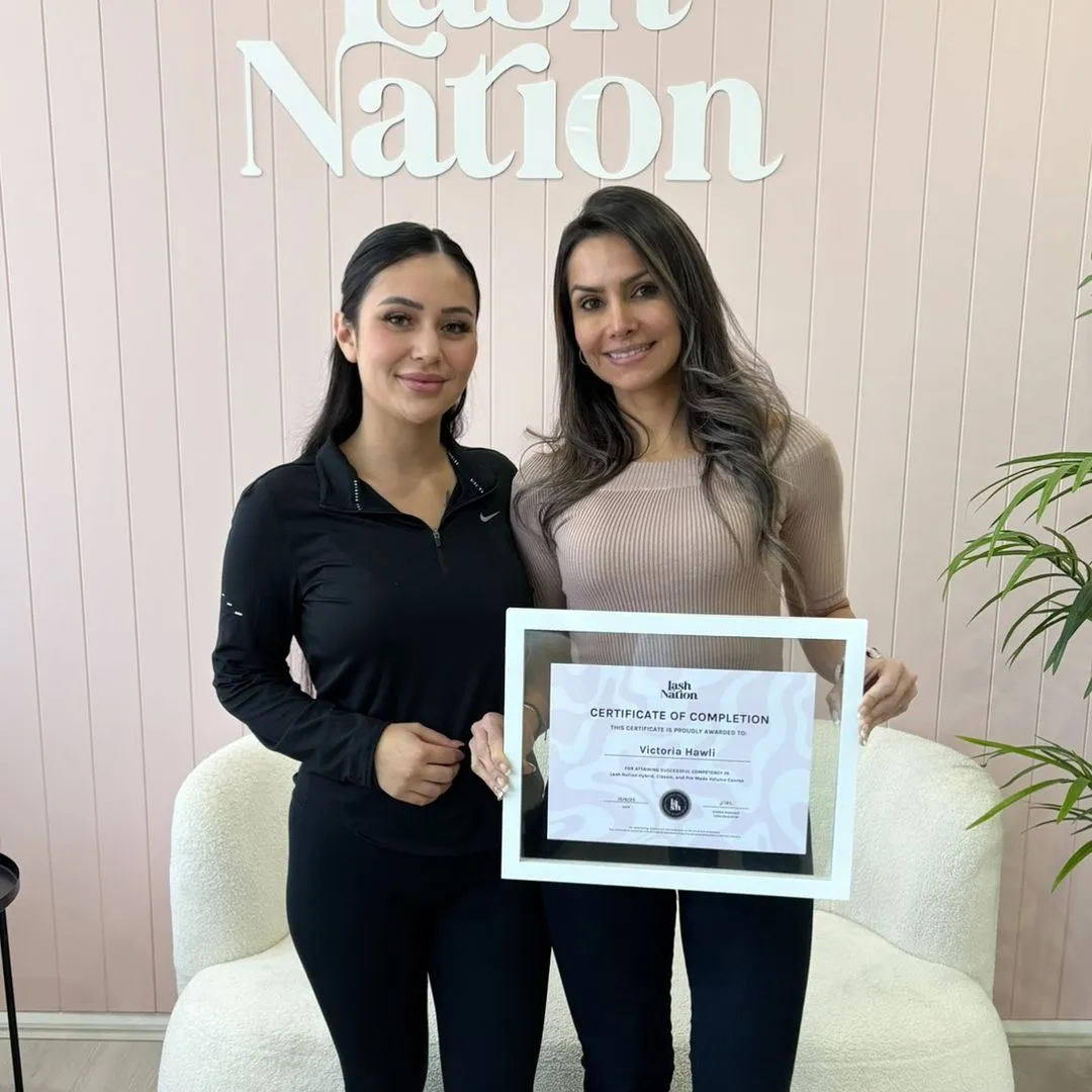 Two women standing indoors holding a framed certificate of completion, with 'Lash Nation' logo visible on a wall behind them.