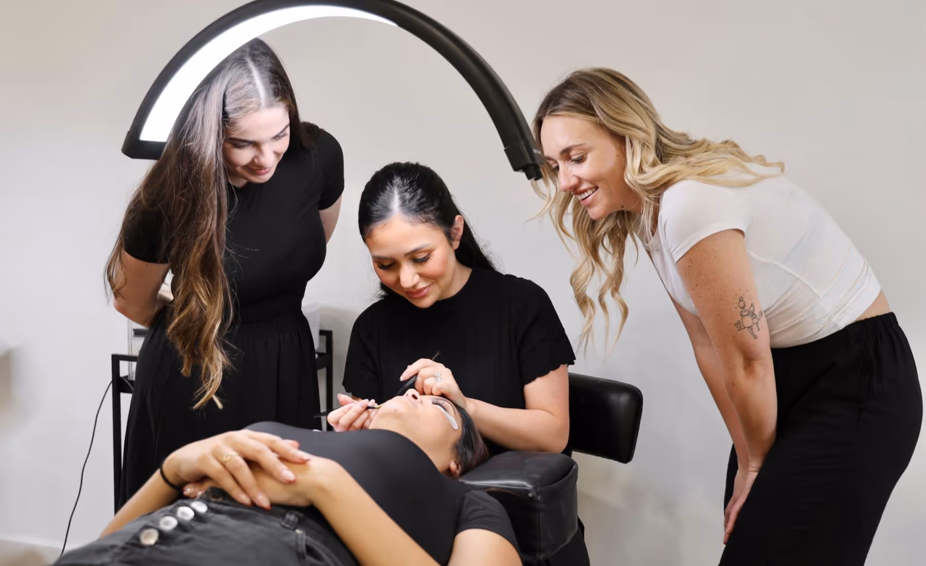 Three women in a beauty salon performing and observing an eyelash extension procedure on a lying client.