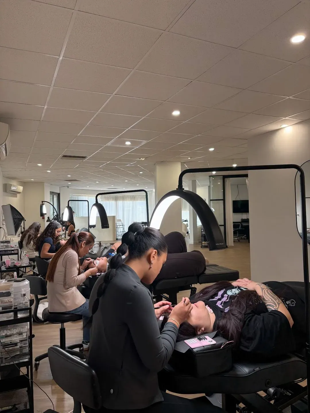 Several beauty technicians apply eyelash extensions to clients lying on black treatment beds under arc-shaped lights in a modern salon.