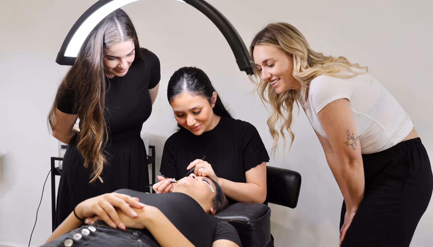 Three women in a beauty salon, one lying down with eye patches while another applies eyelash extensions and a third watches.