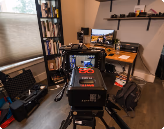 Professional video camera on tripod filming an indoor office with a desk, computer, bookshelf, and camera equipment cases.