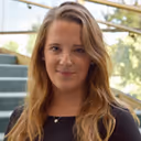 Smiling woman with long light brown hair standing outdoors near stairs and greenery.