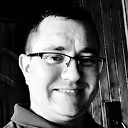 Smiling man wearing glasses in a black and white photo with a wooden background.