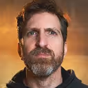 Portrait of a bearded man with medium-length hair against a warm, blurred background.