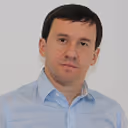 Portrait of a man with short dark hair wearing a light blue collared shirt against a plain pale background.