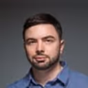 Portrait of a man with short dark hair and a beard wearing a blue collared shirt against a gray background.