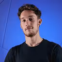 Young man with short curly hair and beard wearing a dark t-shirt against a blue background.