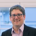 Smiling man with glasses wearing a dark jacket and light purple shirt in an office setting.
