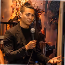 Person in a black blazer speaking into a microphone in a warmly lit indoor setting with artwork in the background.