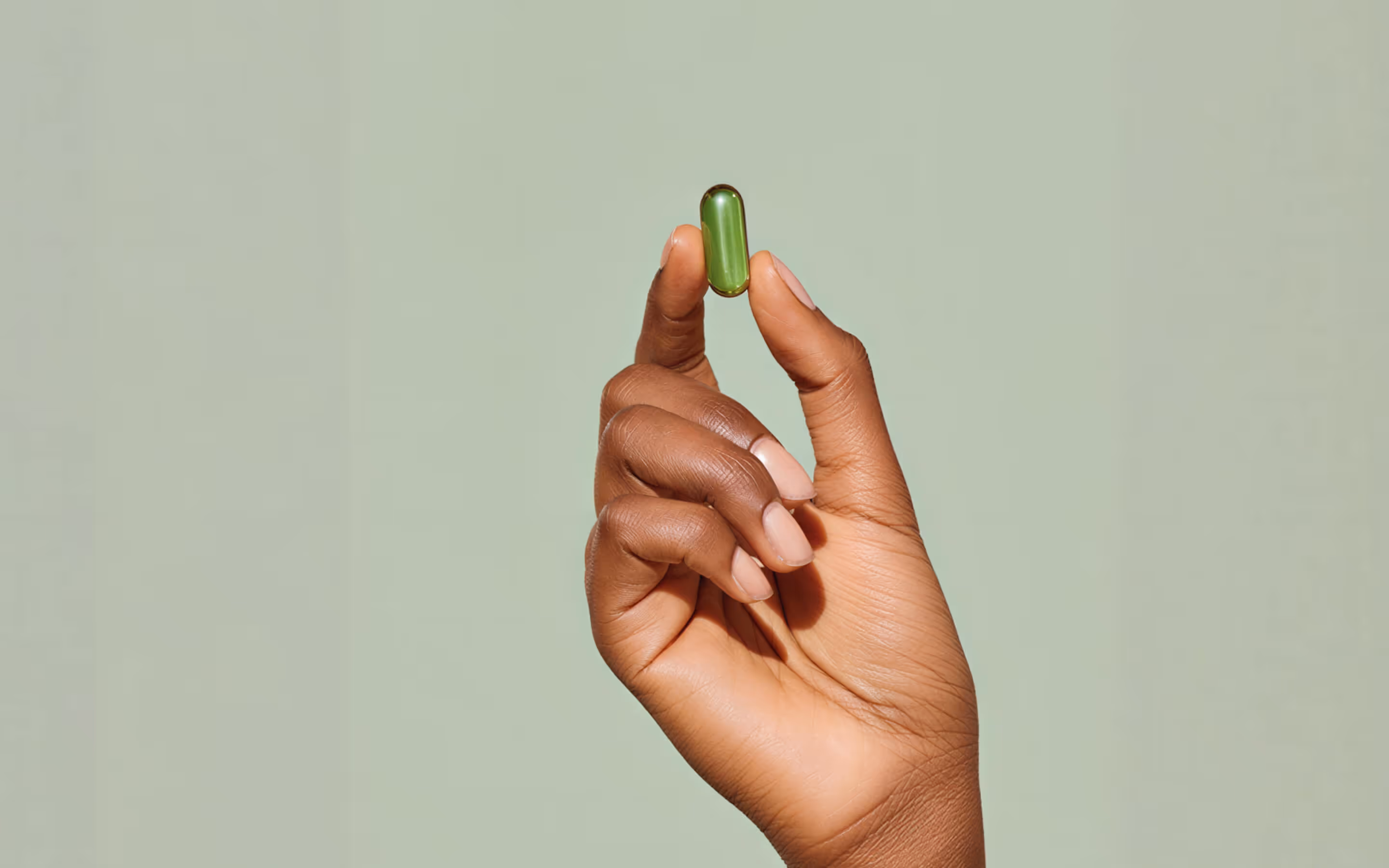 Hand holding a small green capsule between thumb and index finger against a plain light background.