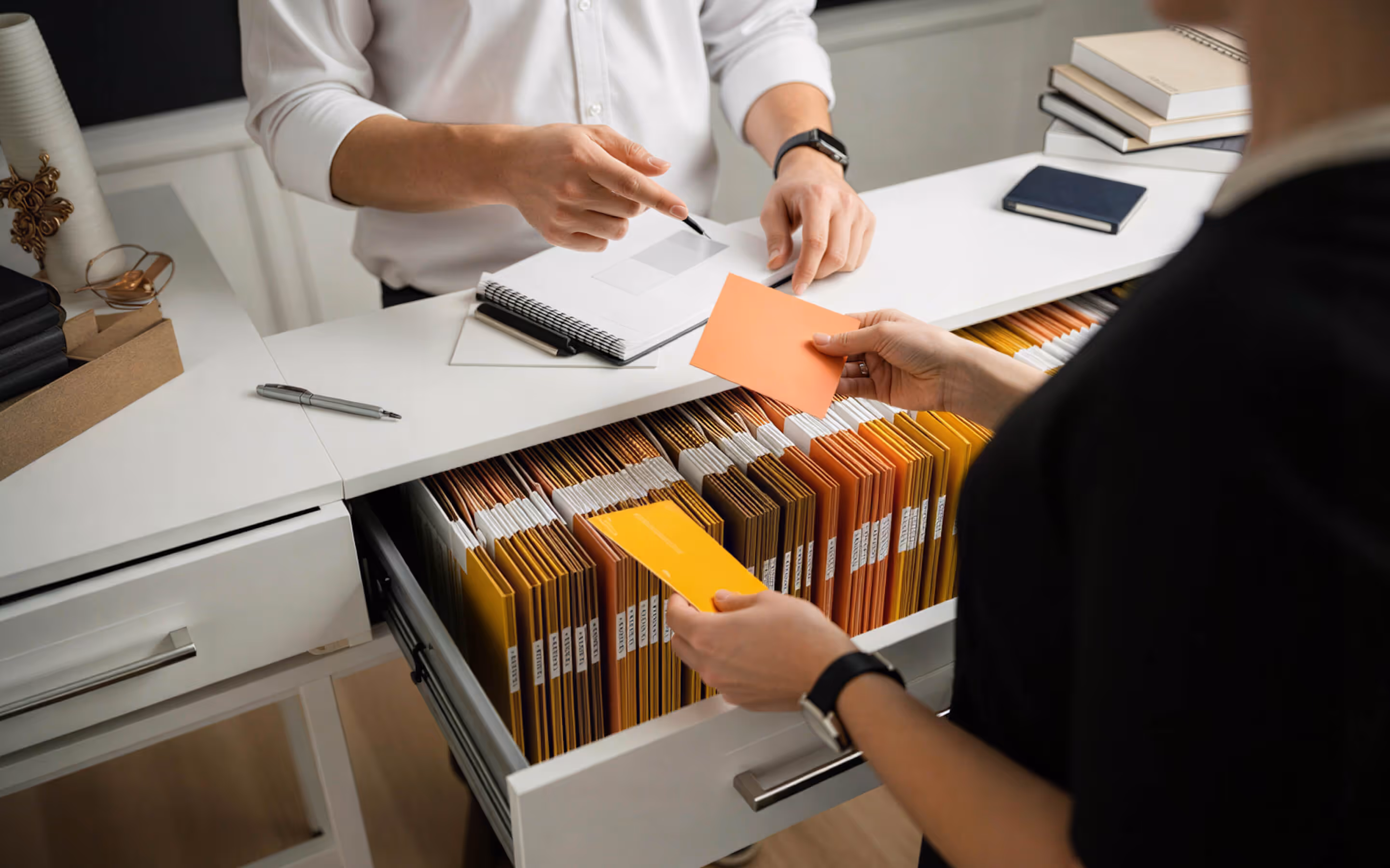 Two people exchanging colored file folders over a desk with an open drawer full of organized documents and notebooks stacked nearby.