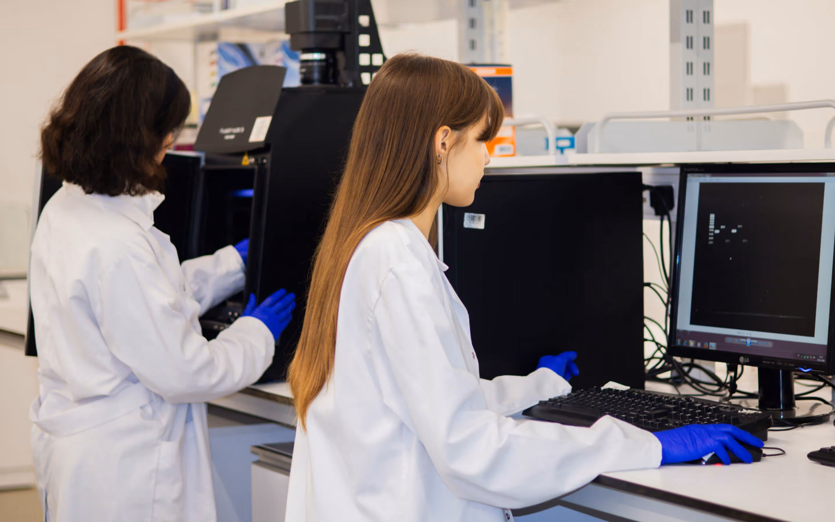Two female scientists in white lab coats and blue gloves working with lab equipment and a computer in a laboratory.