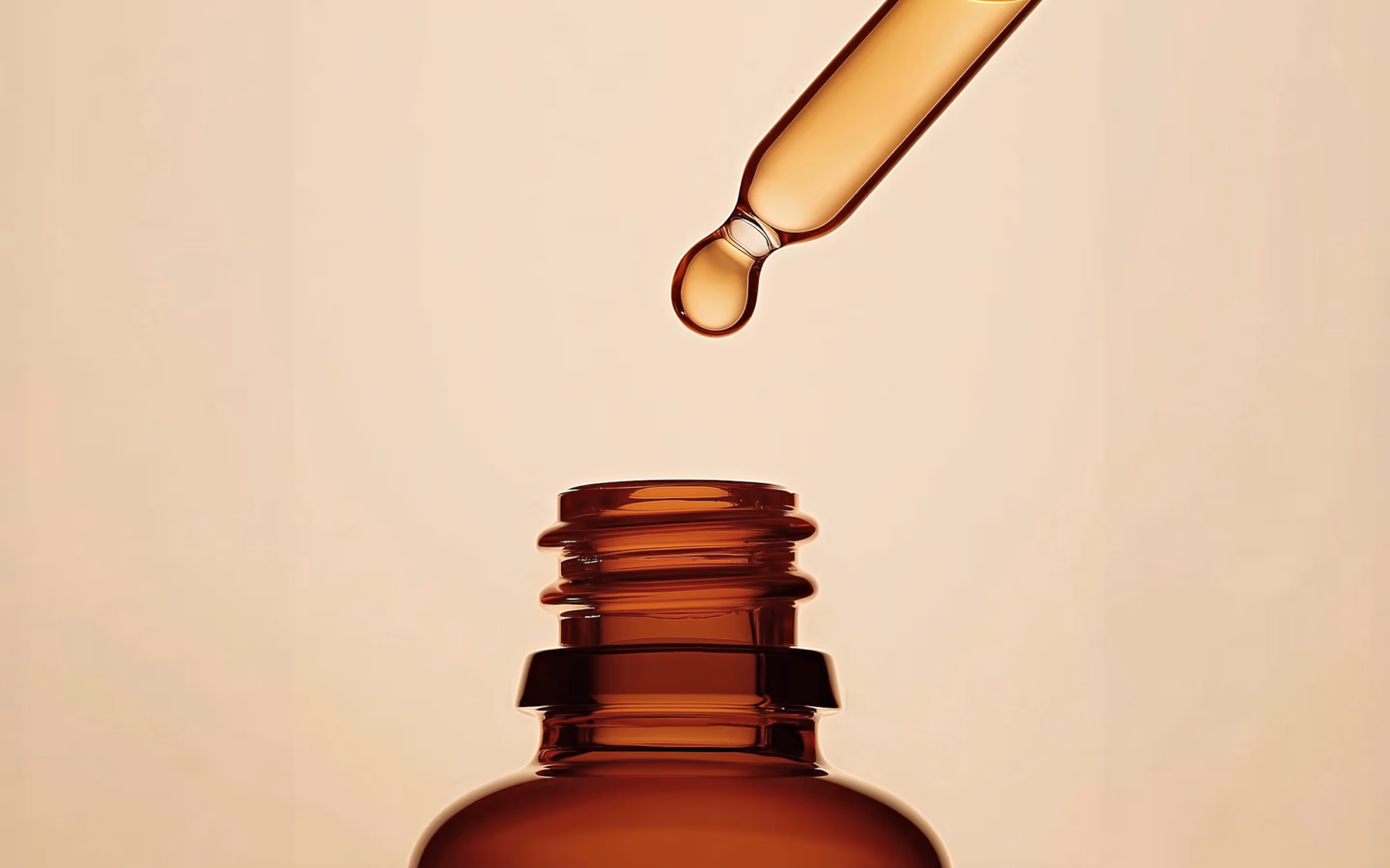 Dropper releasing a drop of liquid into an open amber glass bottle against a beige background.