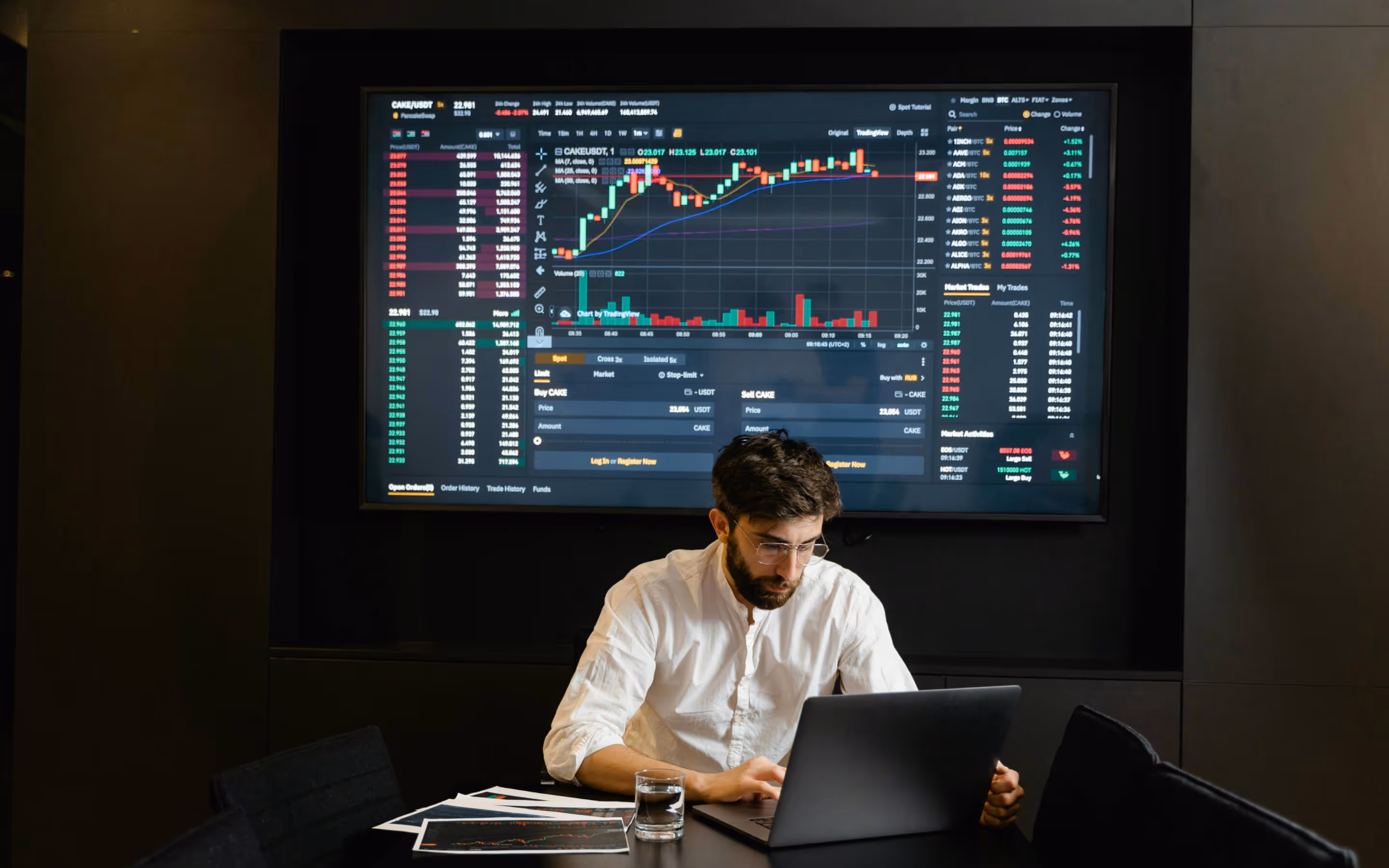 Man in white shirt working on a laptop with financial trading charts displayed on a large screen behind him.