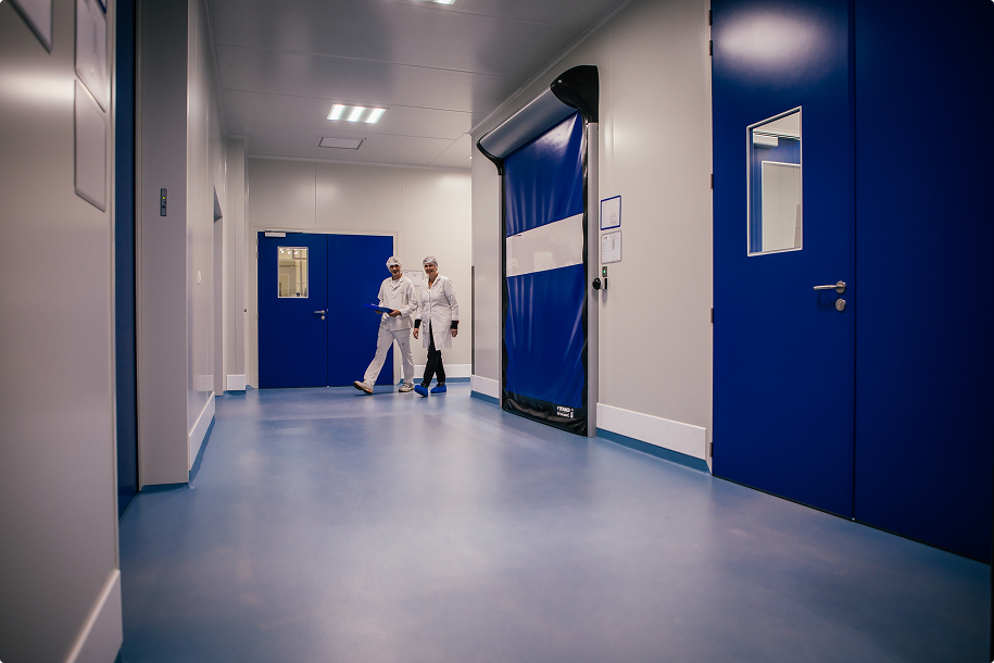 Twee mensen in witte laboratoriumjassen en haarnetjes lopen in een schone gang met blauwe deuren en een automatische blauwe roldeur.
