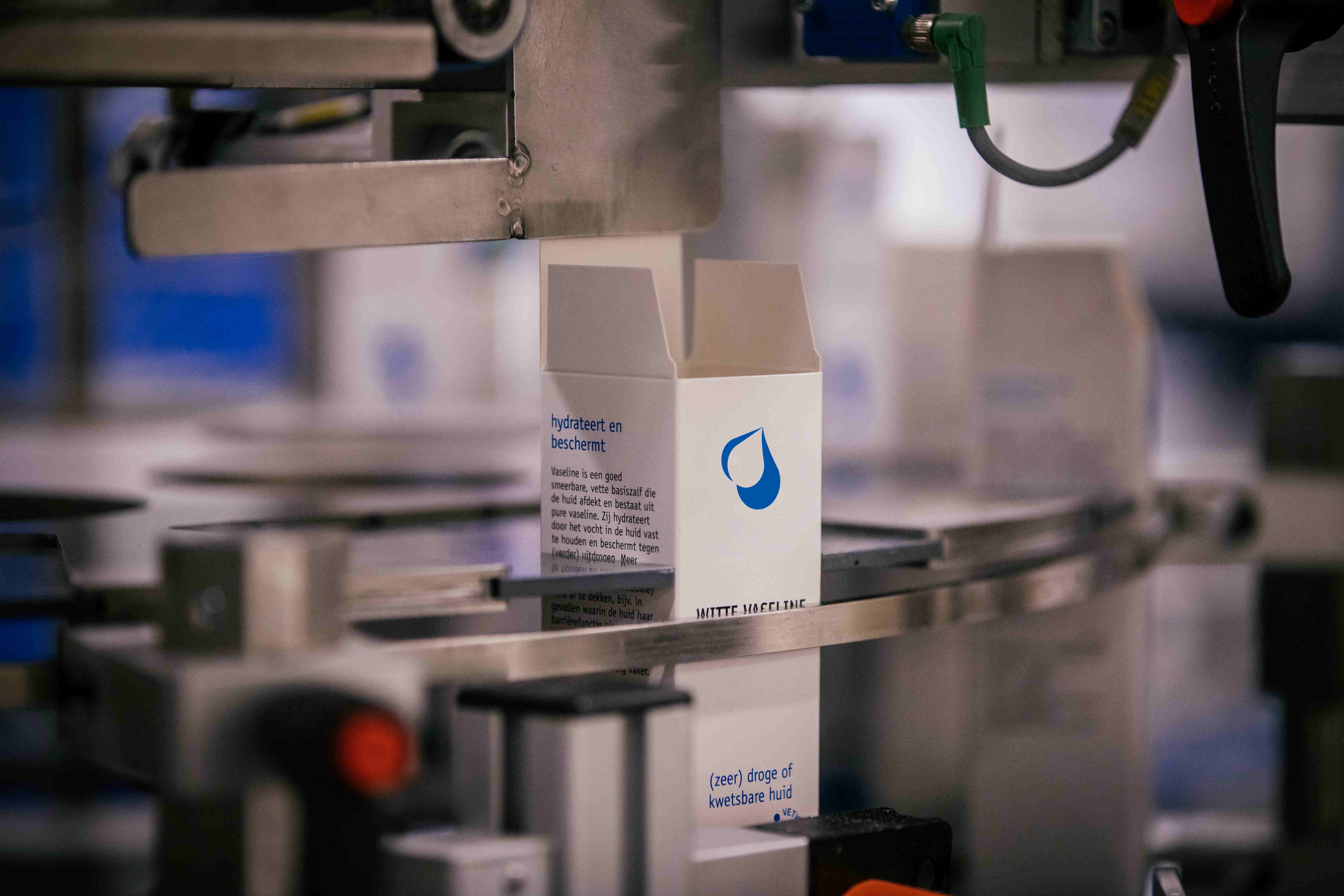 Verpakkingsmachine die een witte doos vaseline met blauwe tekst en druppelicoon op een lopende band verwerkt.