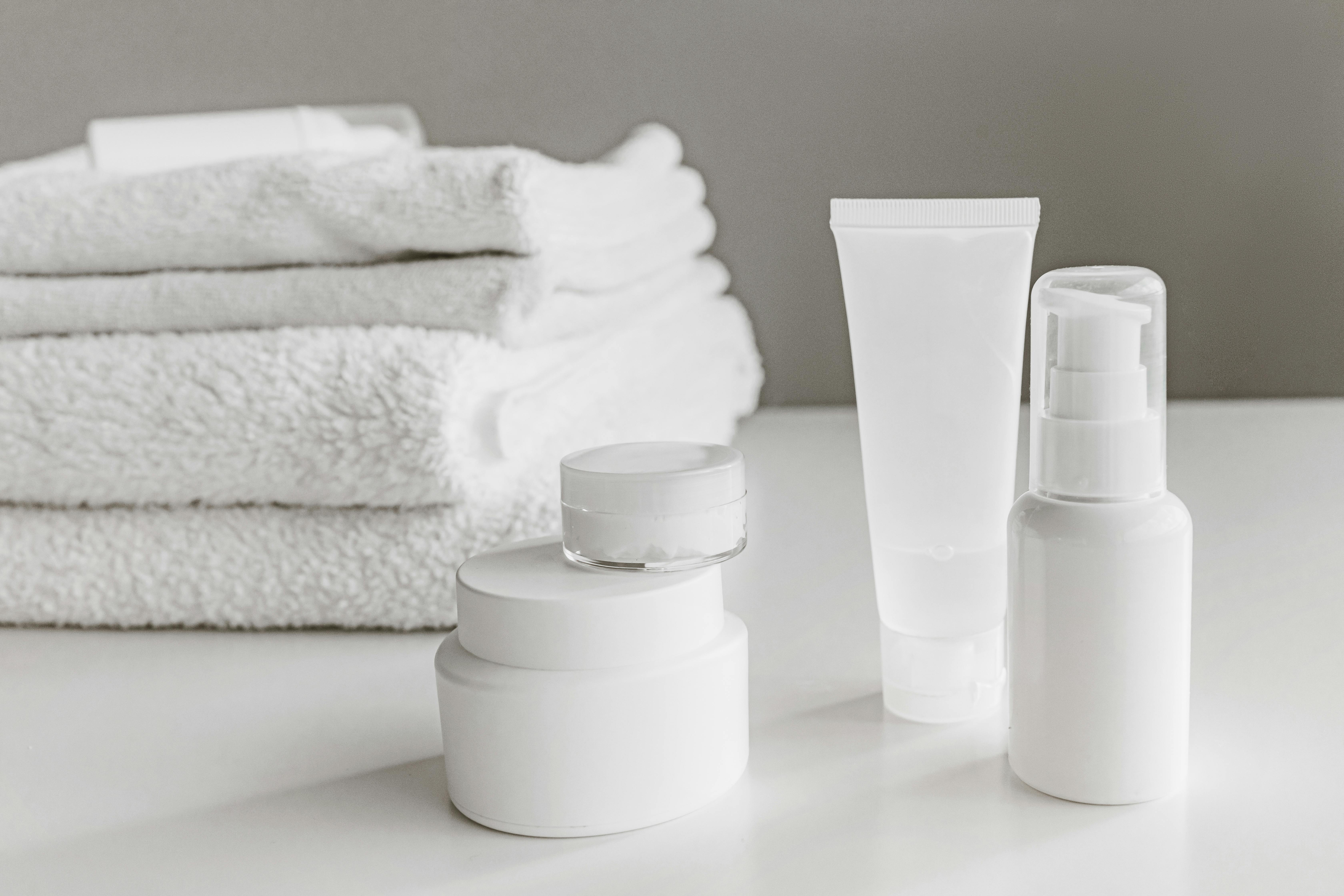 Stapels witte badhanddoeken naast witte cosmetische potjes, tube en doseerpompfles op een witte tafel.