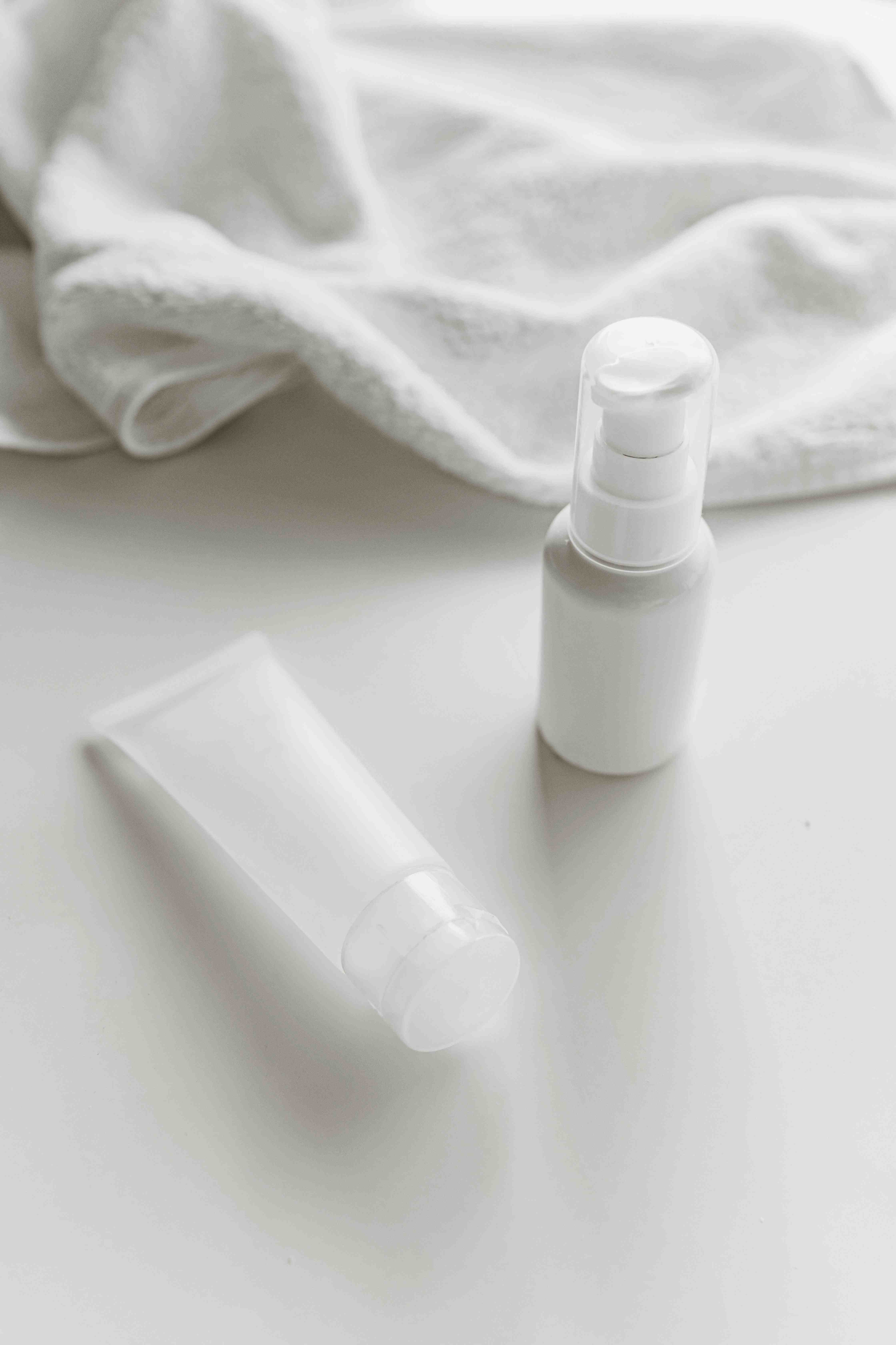 Twee lege witte cosmetische verpakkingen op een witte ondergrond met een witte opgerolde handdoek op de achtergrond.