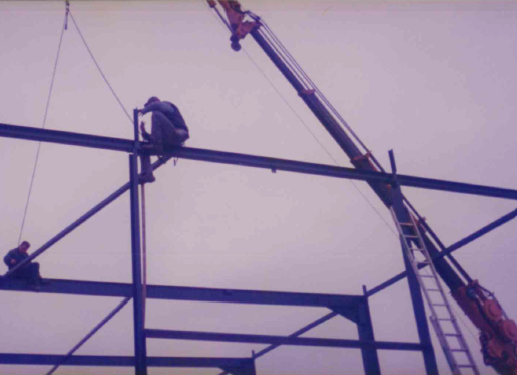 Twee bouwvakkers die op stalen balken werken aan een constructie, met een hijskraan ernaast.