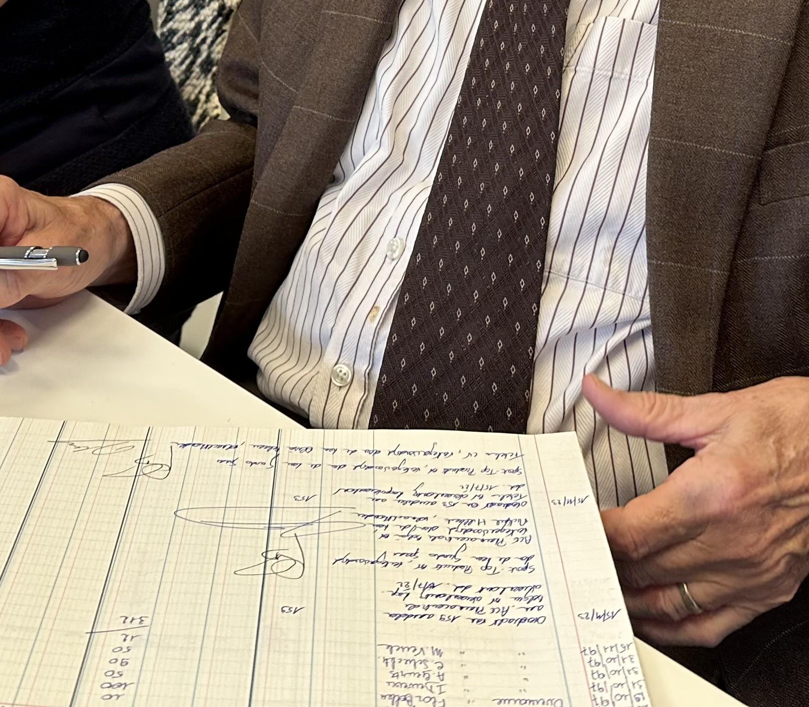 Man in bruine jas en stropdas schrijft aantekeningen op een groot papier met blauwe inkt.