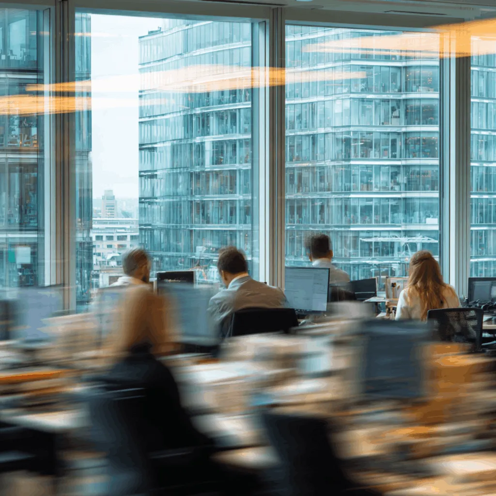 Blurred image of people working in an office with computers and warm lighting.