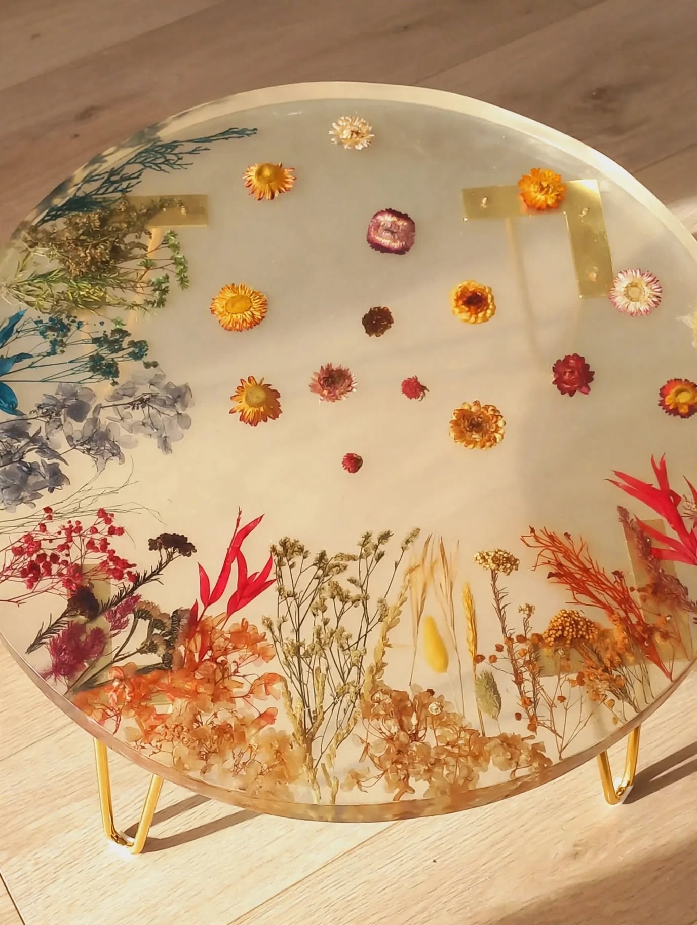 Mesa Bloom 60cm resina transparente con flores secas preservadas sobre pavimento de madera clara natural patas doradas