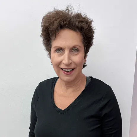 Smiling middle-aged woman with short curly hair wearing a black V-neck top against a light gray background.