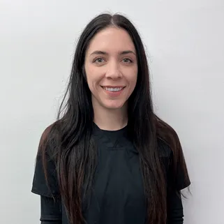 Smiling woman with long dark hair wearing a black shirt standing against a plain light gray background.