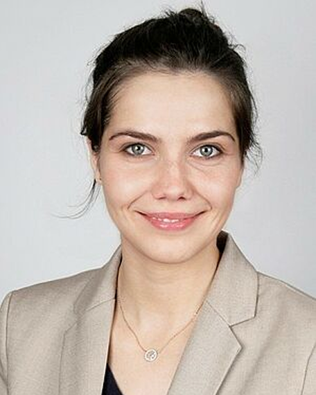 Porträt einer lächelnden Frau mit hochgestecktem dunklem Haar, die einen beigen Blazer und eine Halskette trägt.