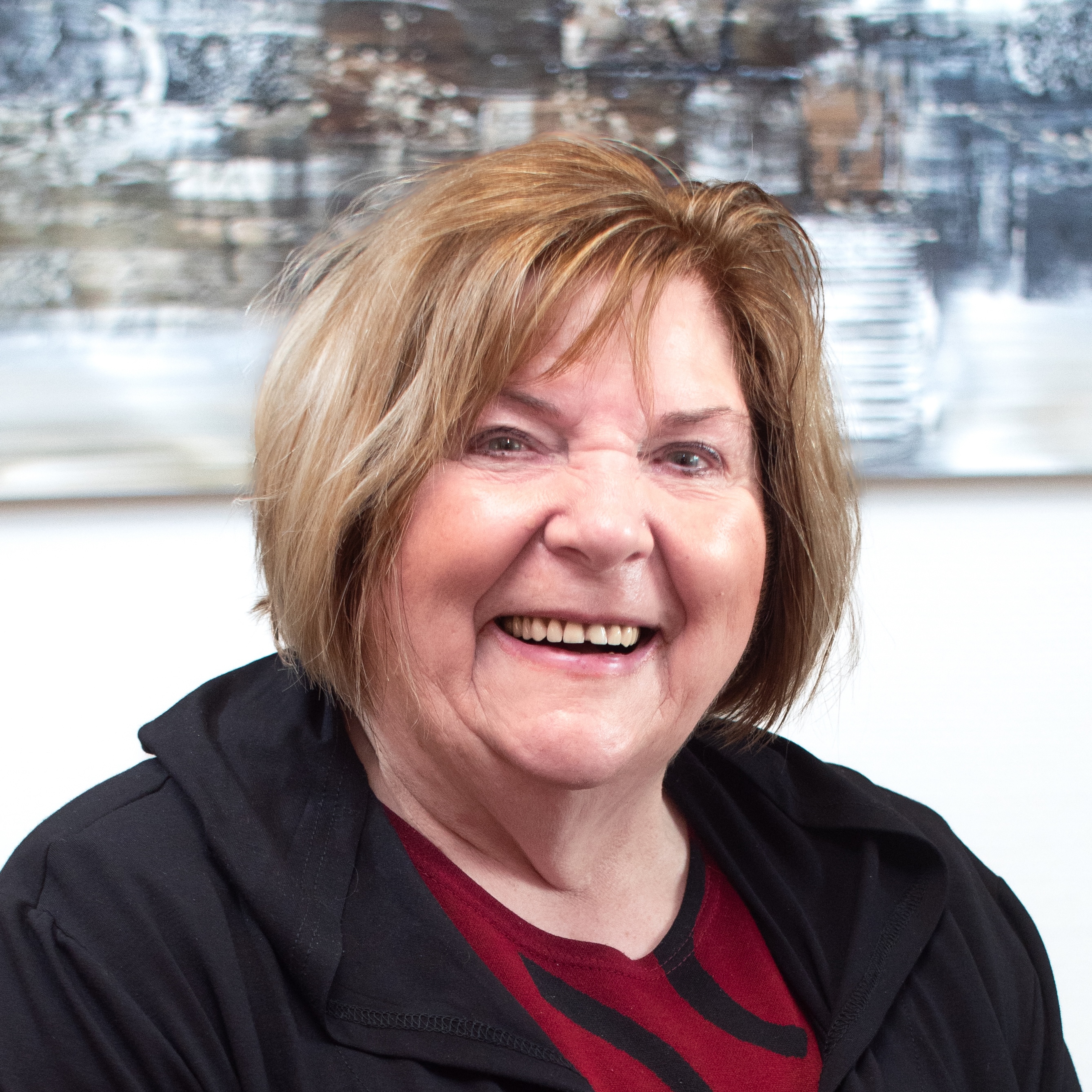 Lächelnde ältere Frau mit kurzen blonden Haaren, die eine schwarze Jacke über einem roten Shirt trägt.