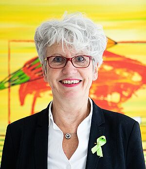 Ältere Frau mit kurzen weißen Haaren, roter Brille, schwarzem Blazer und weißer Bluse vor einem bunten, gelb-roten Hintergrund.