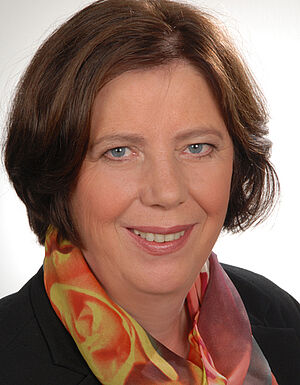 Porträt einer lächelnden Frau mit braunen Haaren, die ein buntes Seidentuch und ein schwarzes Sakko trägt.