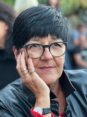 Porträt einer Frau mit kurzen schwarzen Haaren, Brille und schwarzer Jacke, die ihr Gesicht mit der Hand stützt.