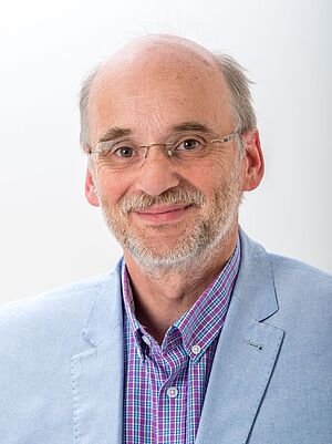 Porträt eines lächelnden Mannes mit Brille, Bart und hellblauem Sakko vor hellem Hintergrund.