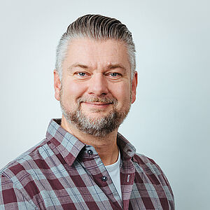 Mittelalterlicher Mann mit grauem und braunem Haar, kurzen Bart, kariertem Hemd und freundlichem Lächeln vor hellem Hintergrund.