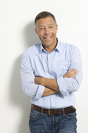 Älterer Mann mit blauem Hemd und dunkler Jeans lehnt lächelnd mit verschränkten Armen an eine weiße Wand.