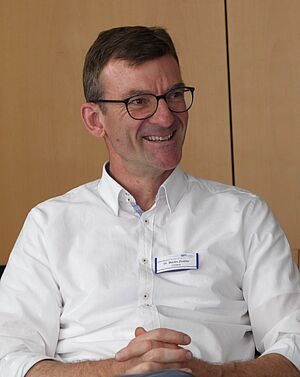 Mann mit kurzen braunen Haaren, schwarzer Brille und weißem Hemd lächelt, vor einer hellbraunen Holzwand sitzend.