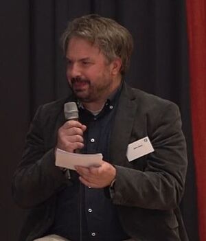 Mann mit Mikrofon und Notizkarte in einem schwarzen Hemd und grauem Sakko vor schwartem Vorhang.