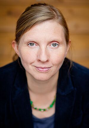 Porträt einer lächelnden Frau mit hellblonden Haaren, blauem Oberteil und grüner Perlenkette vor holzfarbenem Hintergrund.