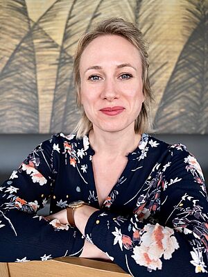 Frau mit blondem Haar trägt ein dunkelblaues, geblümtes Oberteil und lächelt in die Kamera vor einem Wandbild mit Palmenmuster.