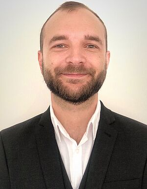 Porträt eines lächelnden Mannes mit kurzem Bart und Anzug vor weißem Hintergrund.