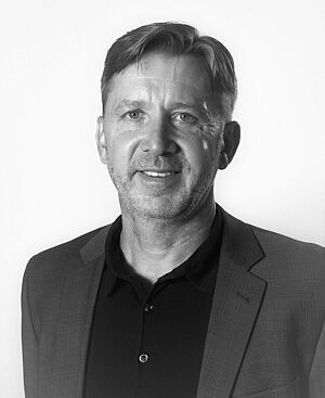 Porträt eines mittleren Mannes mit kurzen Haaren, der einen dunklen Anzug und ein Hemd trägt und in die Kamera lächelt.