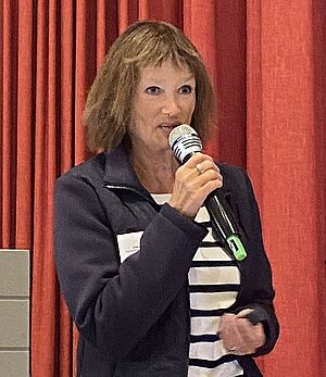 Frau mit mittellangem braunem Haar hält Mikrofon und spricht vor rotem Vorhang.