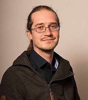 Porträt eines Mannes mit Brille, langen Haaren und dunkelgrauer Jacke vor beigefarbenem Hintergrund.