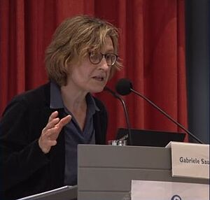 Frau mit kurzen, lockigen Haaren und Brille spricht an einem Rednerpult vor rotem Vorhang.